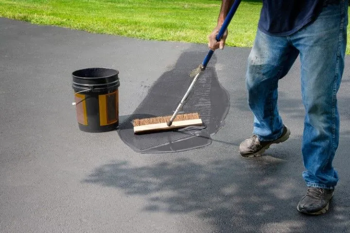 Person applying sealant to a dark driveway with a brush, next to a bucket. Sunny outdoor setting (Benton, Arkansas).