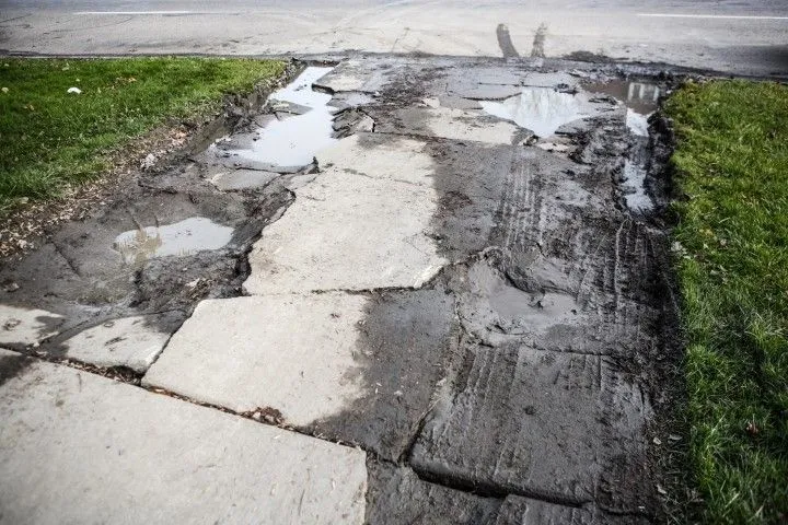 Damaged sidewalk, broken concrete slabs, puddles, mud, and grass alongside a road (Benton, Arkansas).
