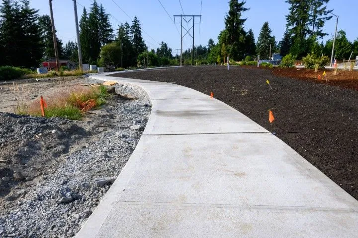 A newly paved concrete sidewalk curves through a park with dark soil and trees in Benton, Arkansas
